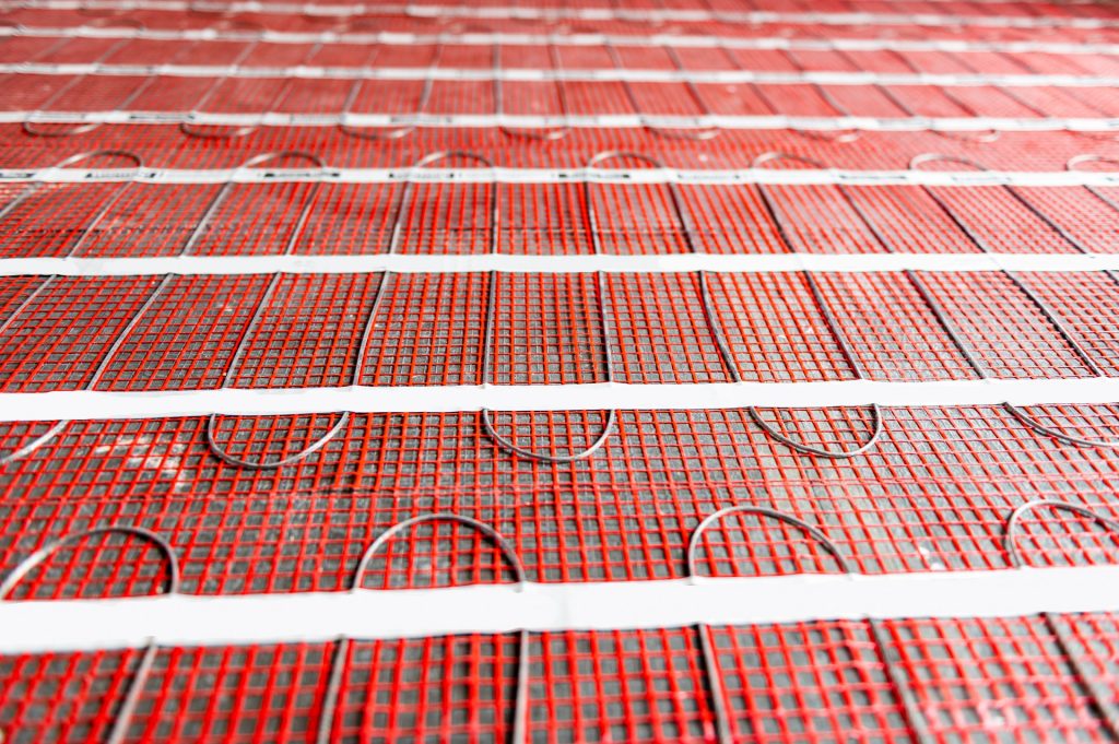 Electric radiant floor heating mats laid in runs across a bathroom subfloor with the heating cable visible in the red mesh before tile goes down
