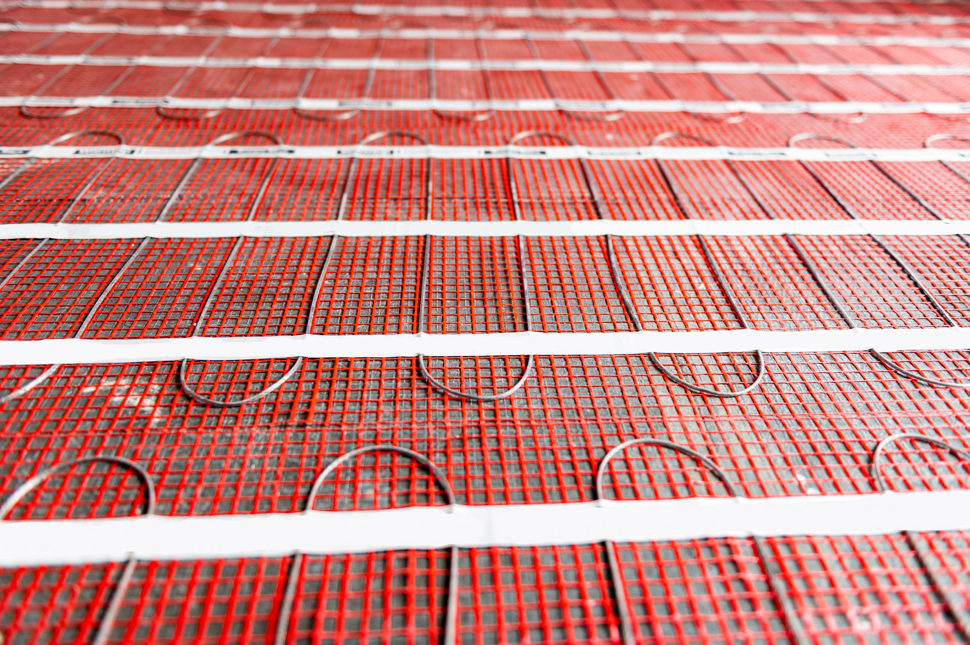 Electric radiant floor heating mats laid in runs across a bathroom subfloor with the heating cable visible in the red mesh before tile goes down