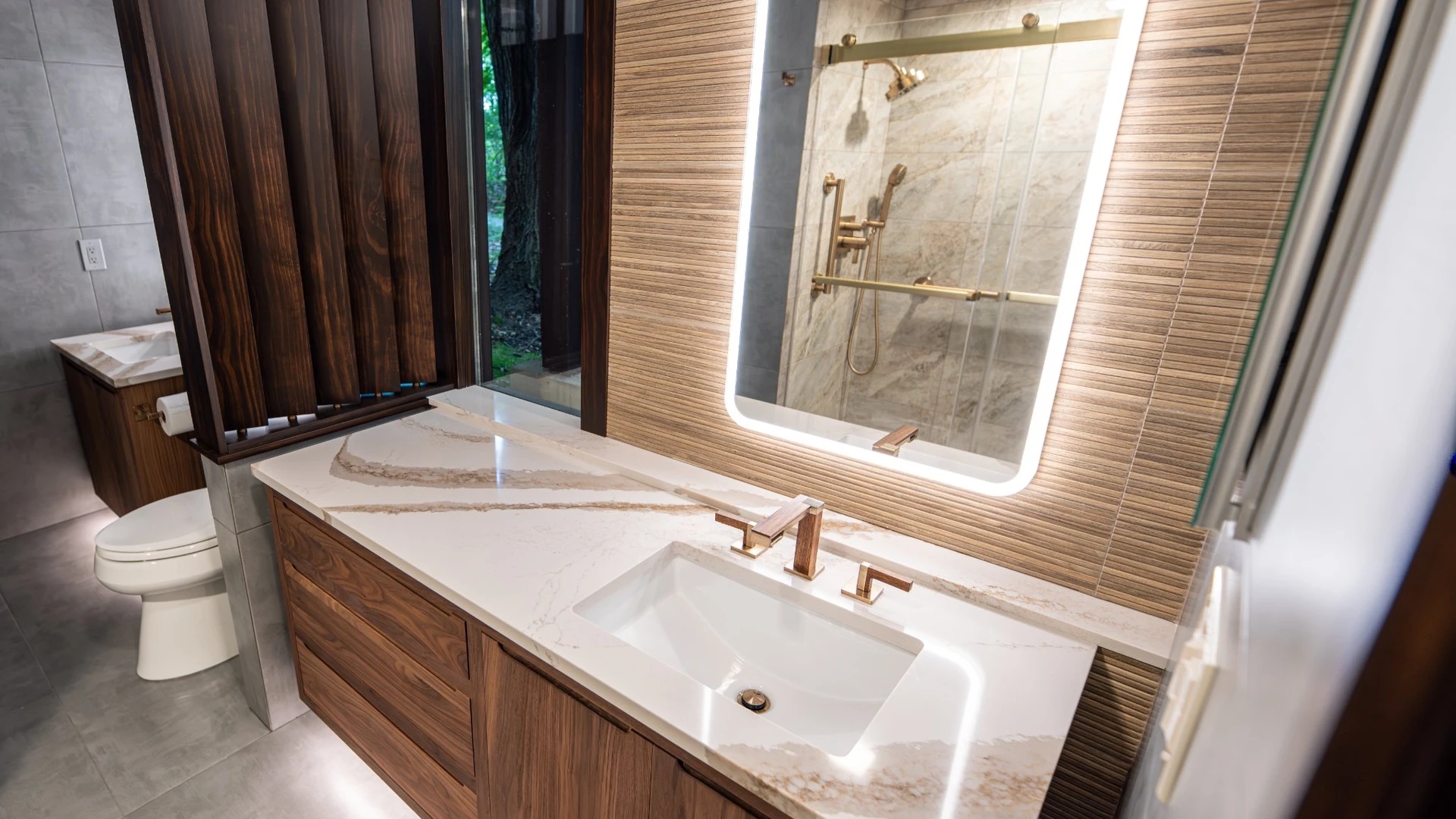 Floating wood vanity with a veined white countertop, gold-tone faucet and backlit mirror reflecting a glass shower