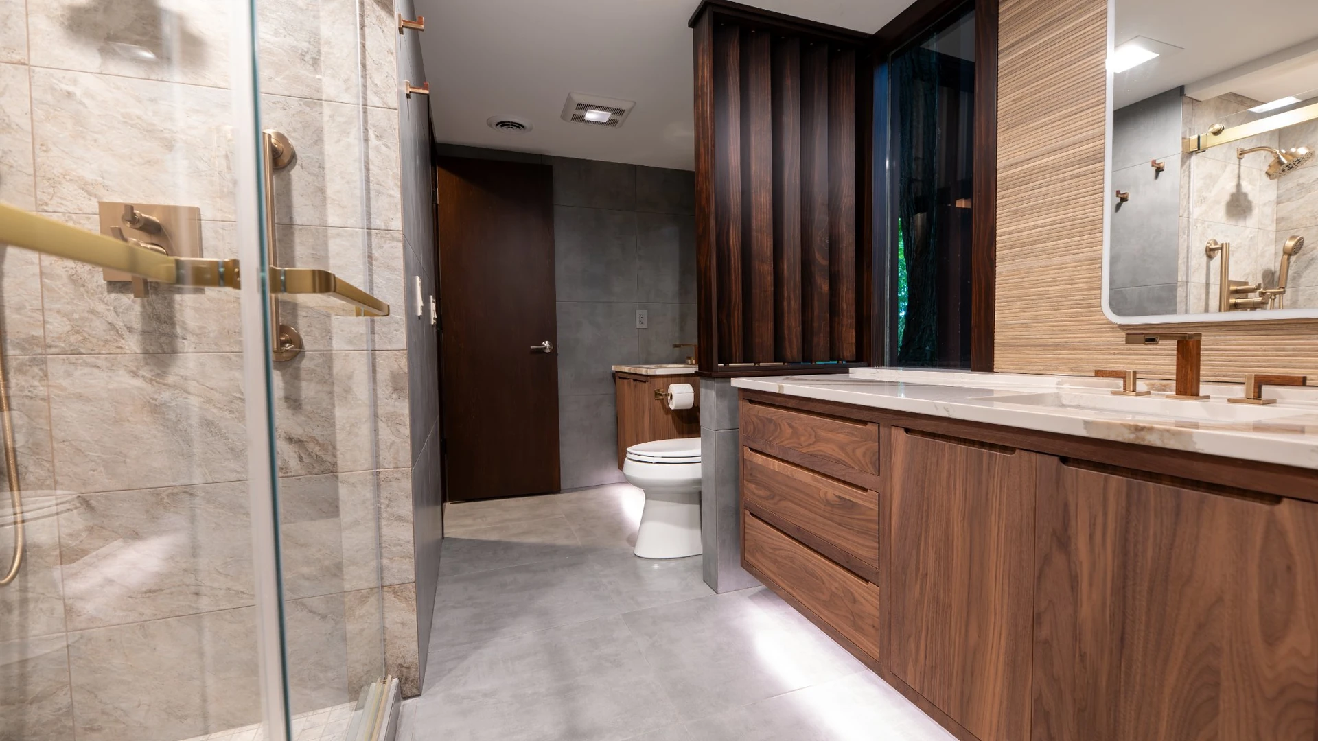 Remodeled bathroom with large-format gray floor tile running from the glass shower past a floating walnut vanity with under-cabinet lighting
