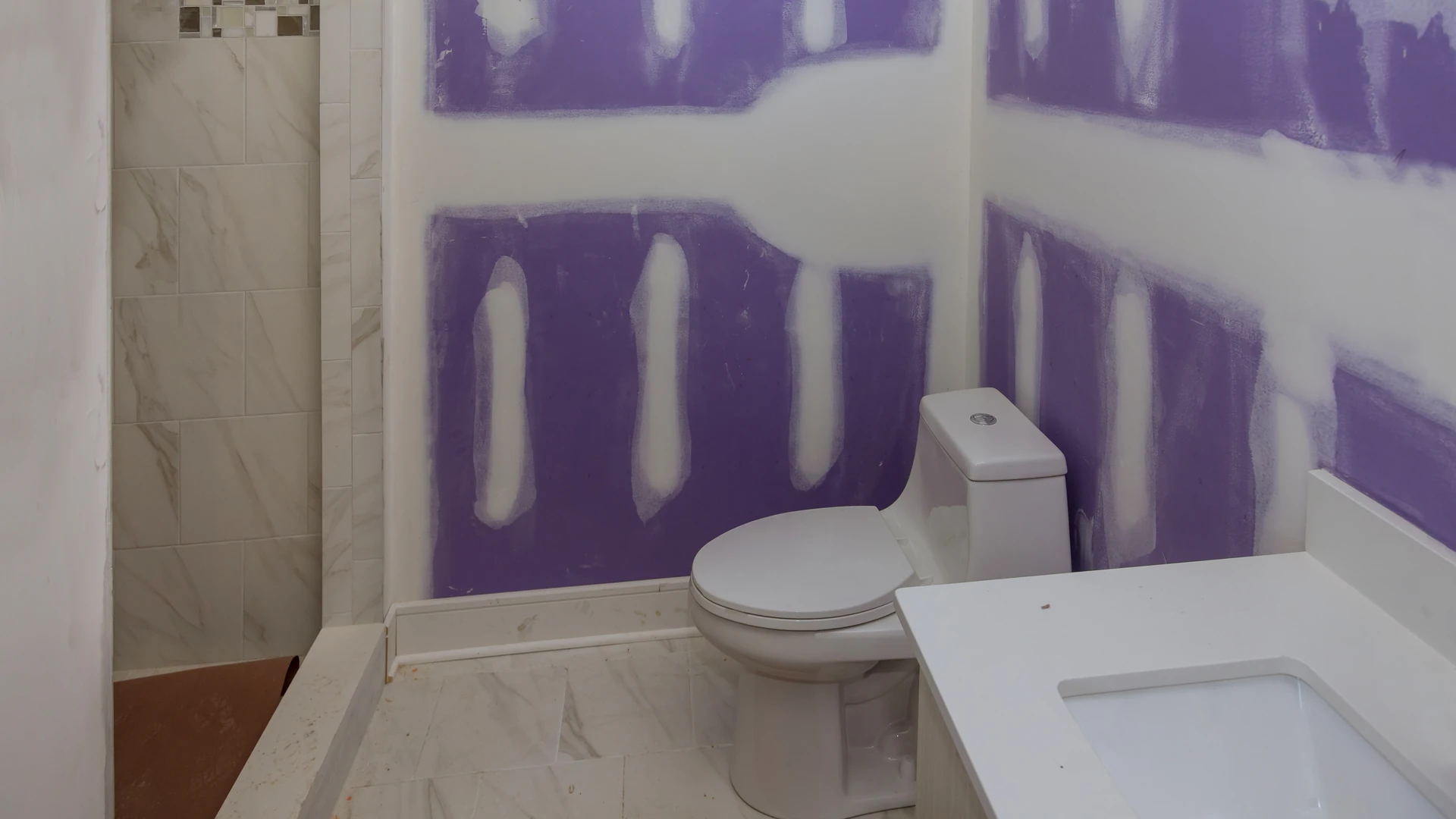 Bathroom mid-remodel with patched purple drywall, a newly set toilet, white vanity and a tiled walk-in shower