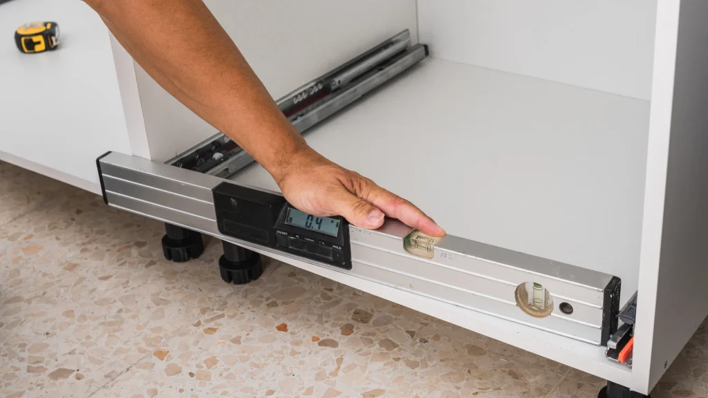A hand adjusts a digital level on a cabinet leveling leg in the empty toe-kick recess beneath a white kitchen cabinet base, before the toe-kick panel is installed
