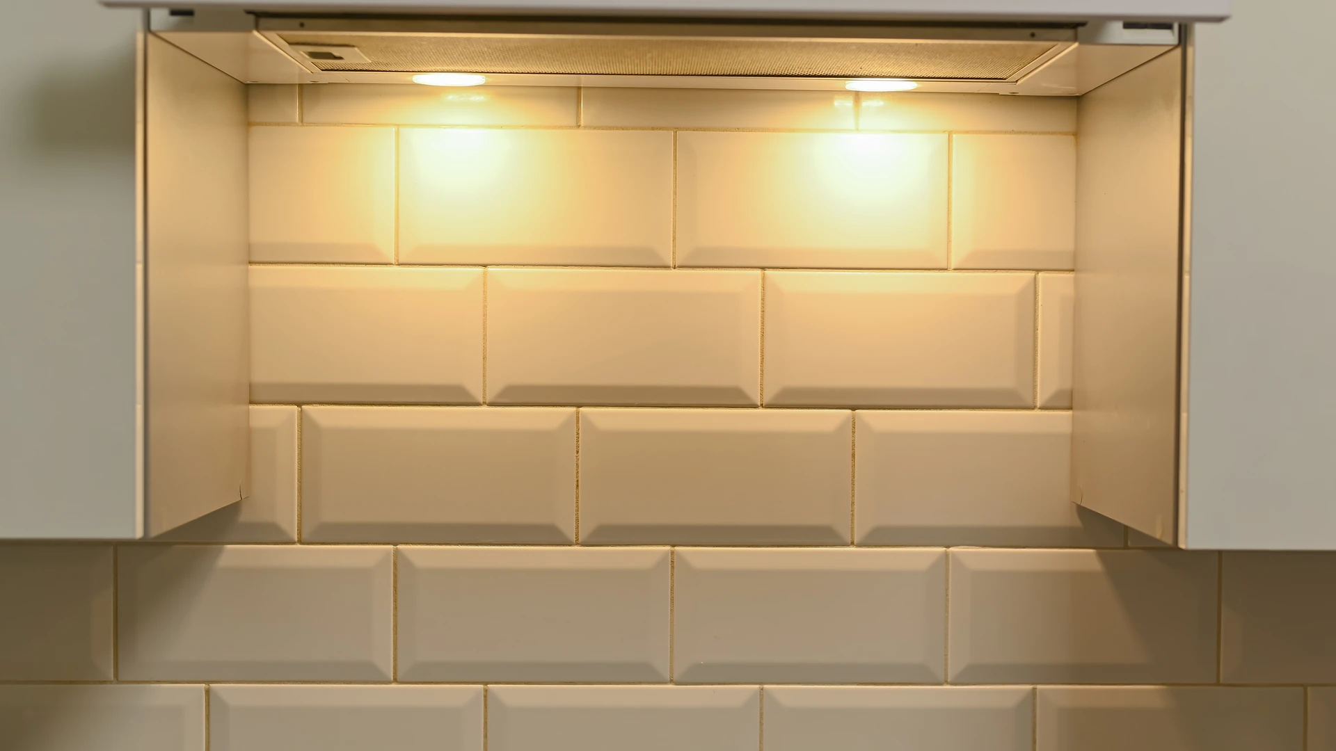 Two warm lights beneath a range hood illuminate a white beveled tile backsplash.