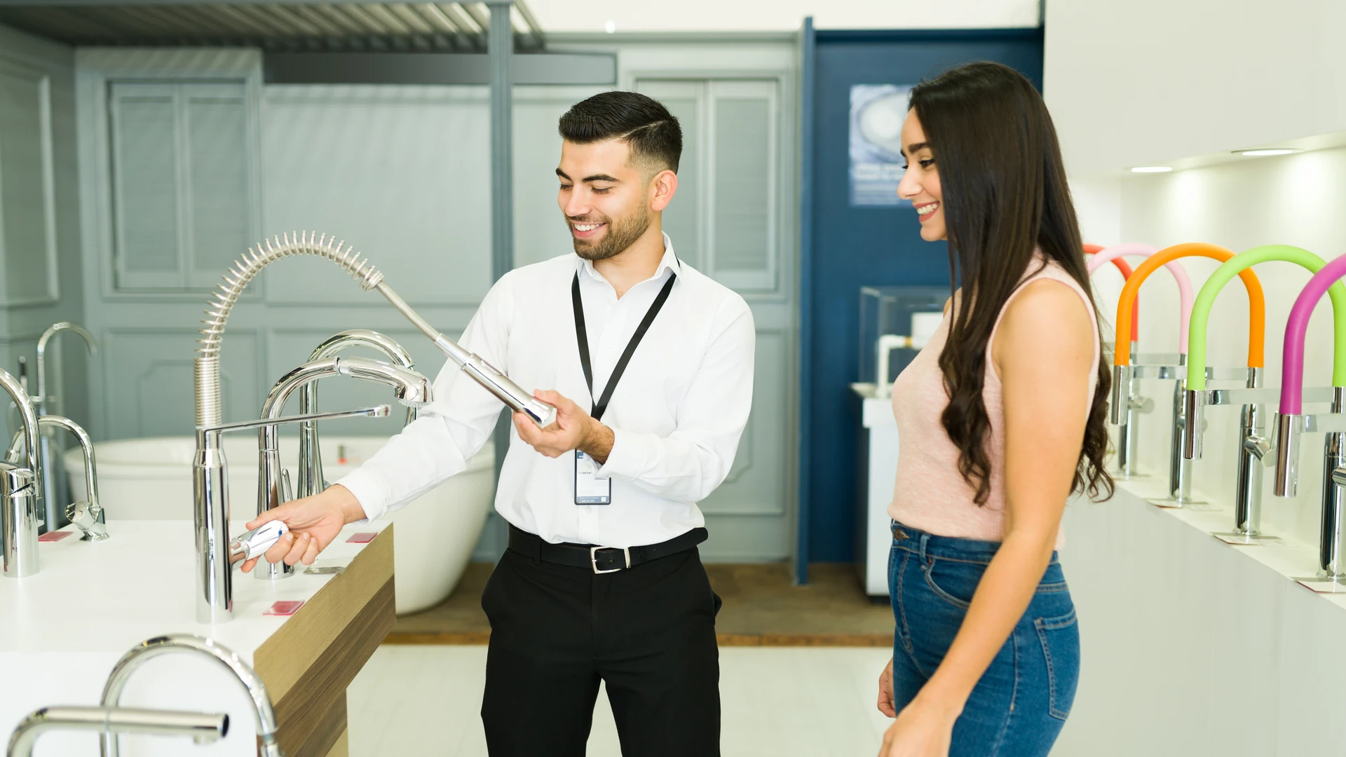 Showroom salesperson demonstrating a pull-down kitchen faucet sprayer to a smiling customer among faucet displays