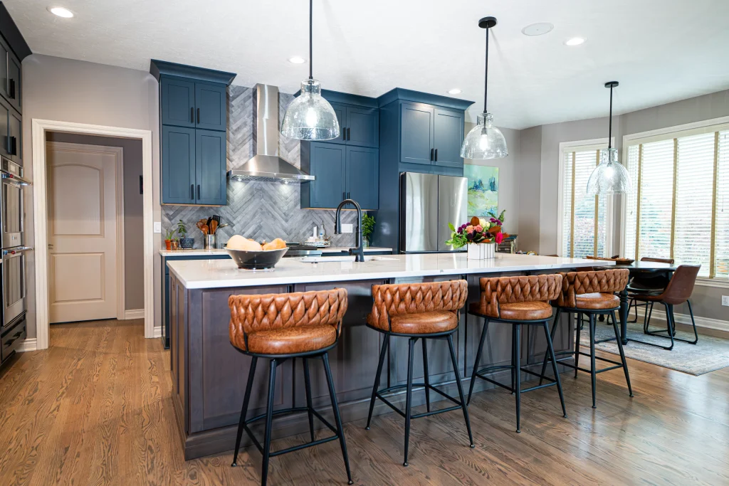 Blue kitchen cabinets behind a broad island with brown stools and glass pendant lights