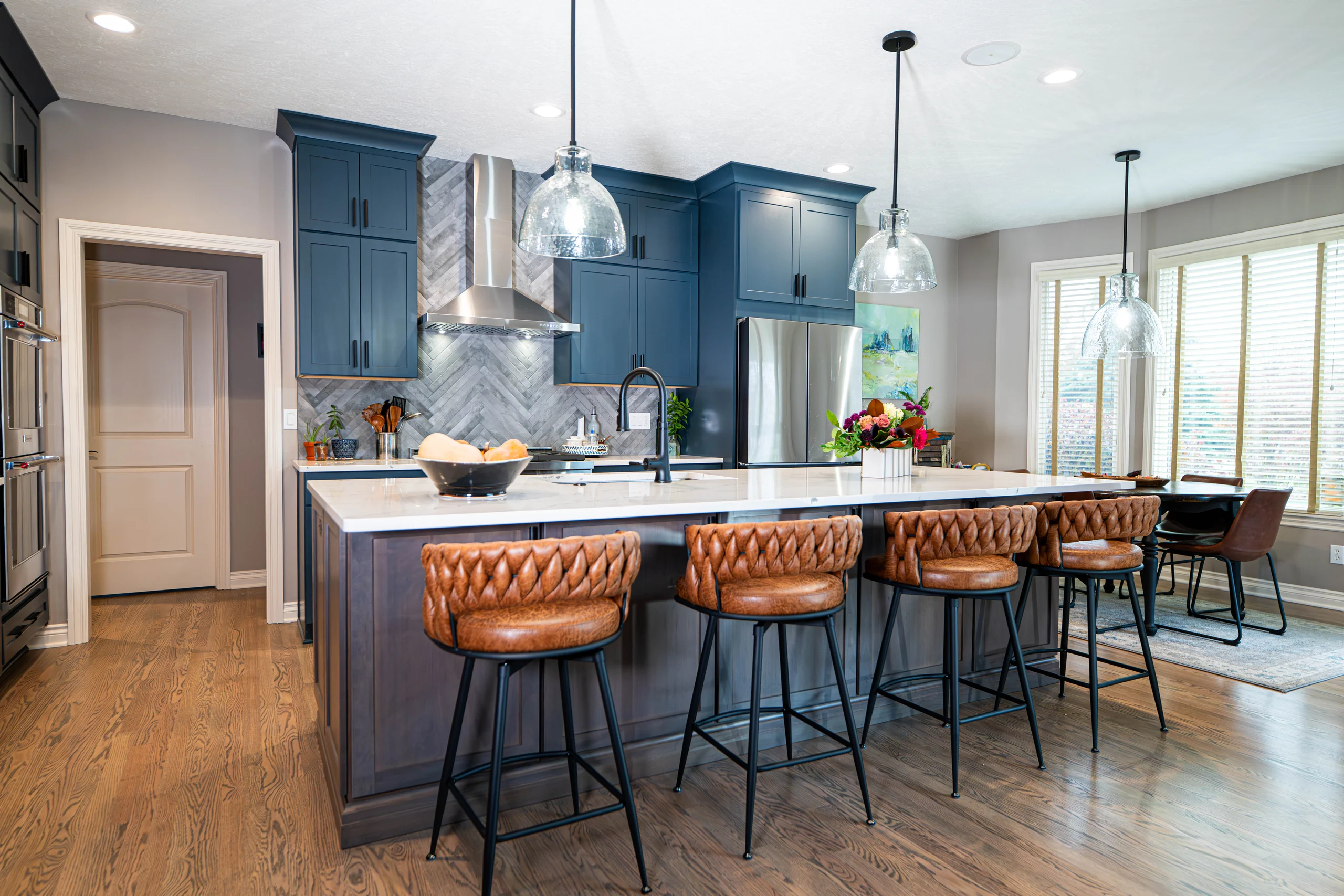 Blue kitchen cabinets behind a broad island with brown stools and glass pendant lights