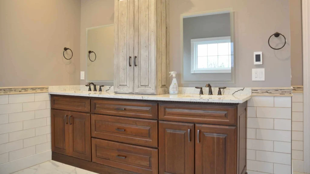 Wood bathroom vanity with two sinks: a drawer-height panel above two doors under each sink and a stack of three drawers between them, with a tall cream cabinet standing on the white veined counter.