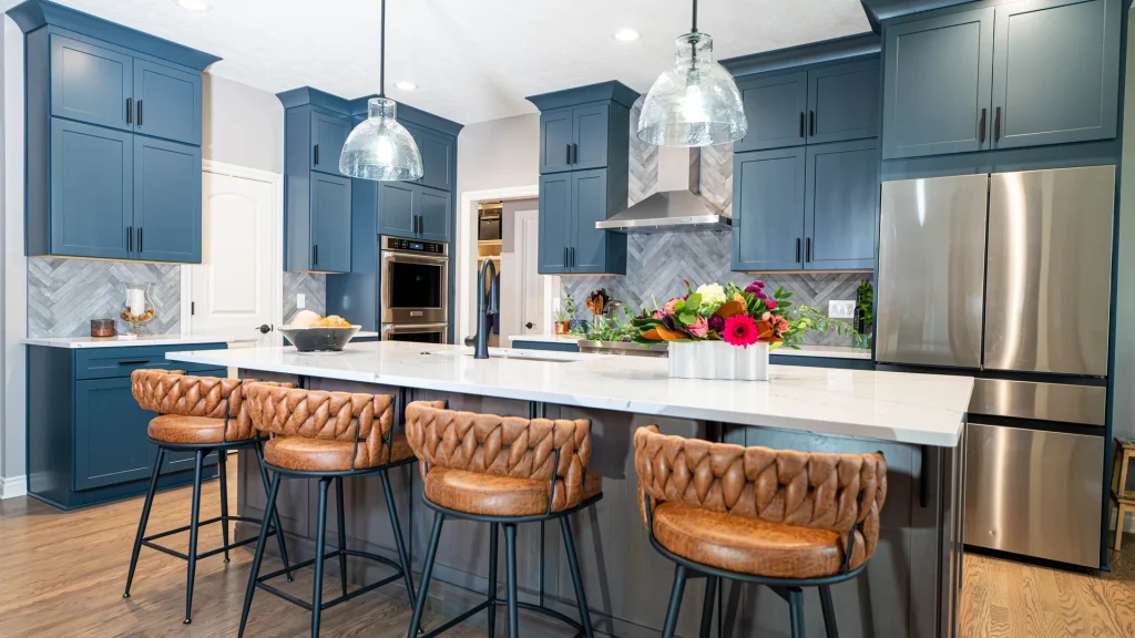 Kitchen with blue shaker-style cabinets, a white-topped island with four woven-back stools, two glass pendant lights, a gray herringbone tile backsplash and a stainless steel refrigerator