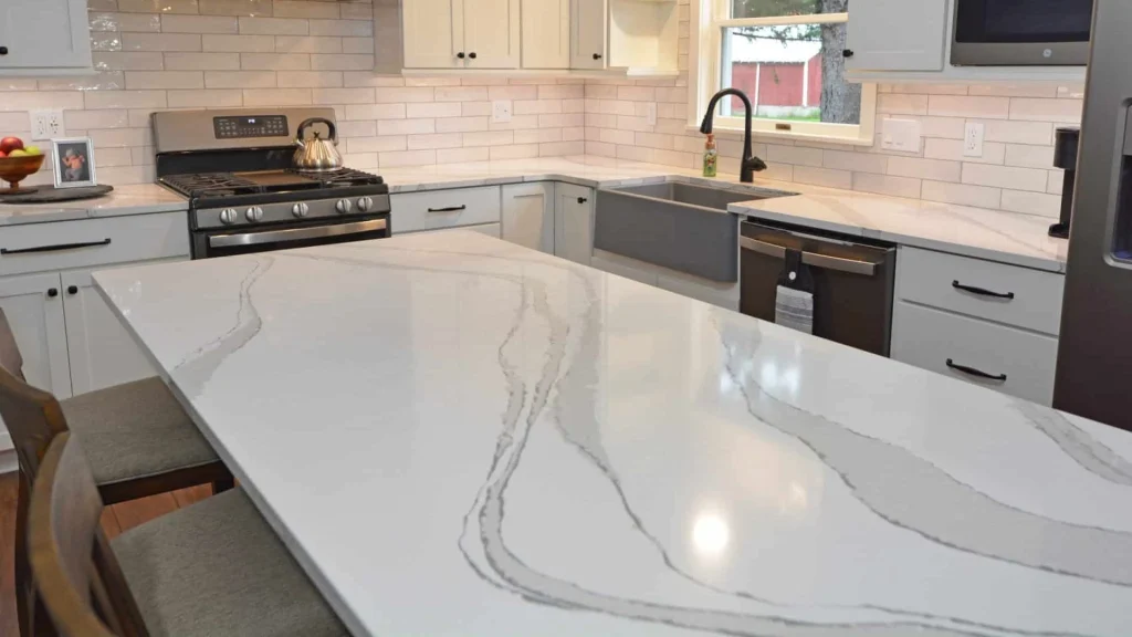 A kitchen with a gray apron-front sink and black faucet by a window, a dishwasher beside the sink and a veined white island counter in the foreground