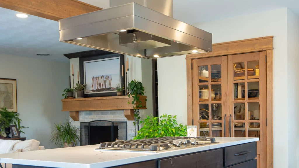 Gas cooktop on a white-topped island beneath a broad suspended stainless steel range hood, with a brick fireplace and glass-door pantry cabinets behind