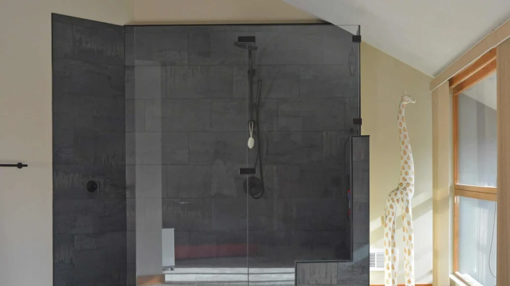 Walk-in shower with dark gray tiled walls and a clear glass panel, a rain shower head on a wall arm and a handheld shower on a slide bar, in a bathroom with a sloped ceiling and a tall window