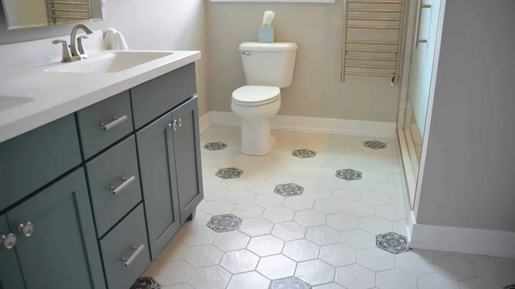 White two-piece toilet against the back wall of a bathroom, beside a dark gray-green vanity and a white hexagon tile floor with dark patterned accent tiles