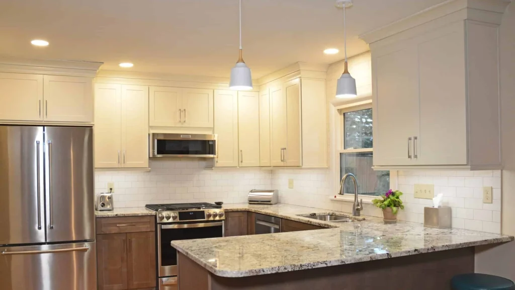 Kitchen with cream upper cabinets, warm brown lower cabinets, a speckled light counter peninsula, white subway tile, two white pendant lights and a stainless steel refrigerator and range