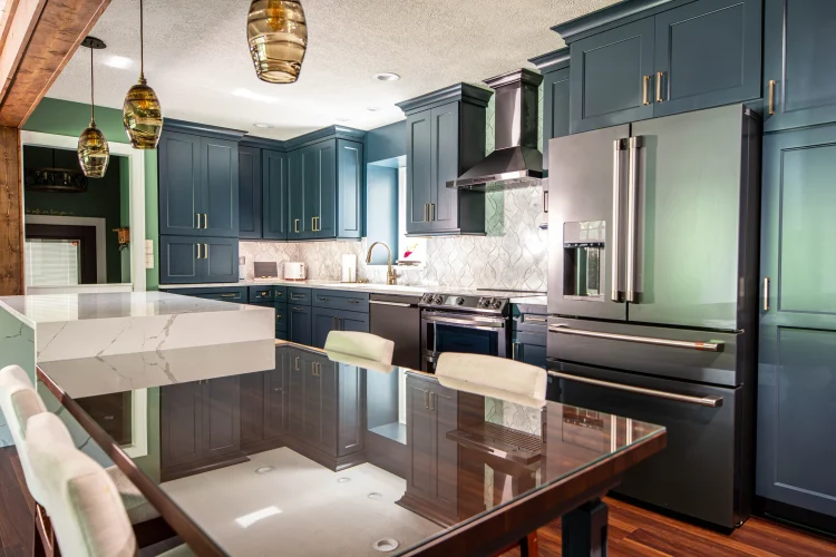 Blue kitchen cabinets and metallic-finish appliances behind a glass-topped dining table with pale chairs